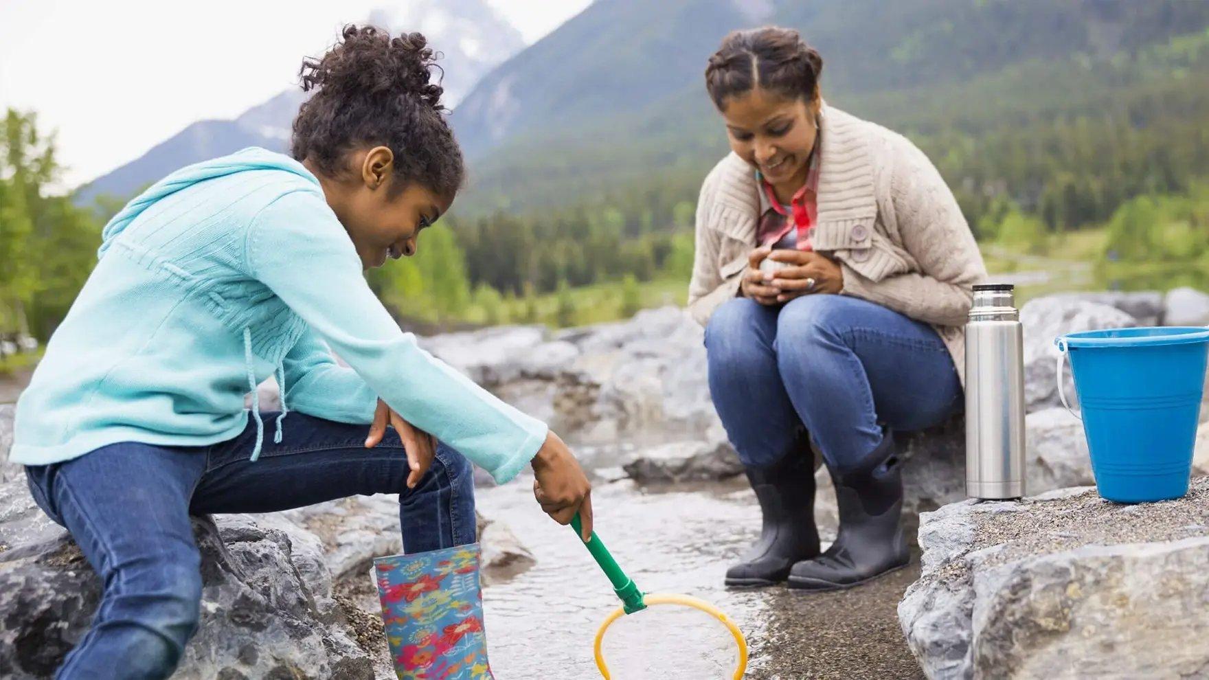 Mujer y niña con chompas y botas de lluvia usando una red para atrapar bichos en un riachuelo poco profundo